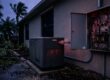 A residential standby generator runs beside a dark house with an open electrical panel showing a tripped breaker.